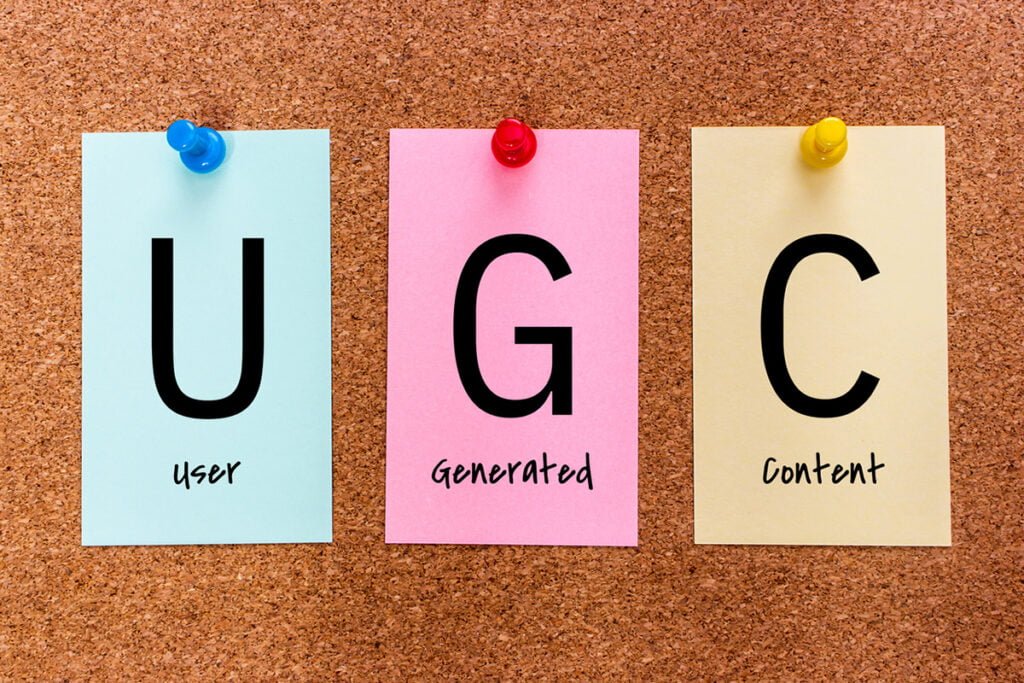 3 letters keyword UGC (User Generated Content) on multicolored stickers attached to a cork board.